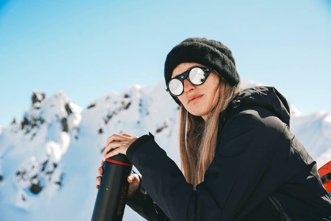 A woman wearing sunglasses and a hat is sitting on top of a snowy mountain.